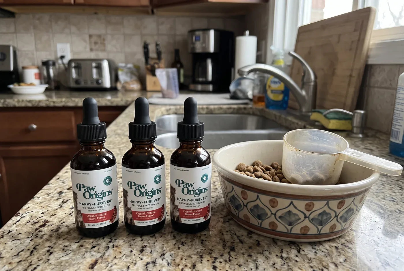 Product shot: three Happy-Furever® full-spectrum CBD oil bottles with droppers on a light wood surface beside a dog bowl