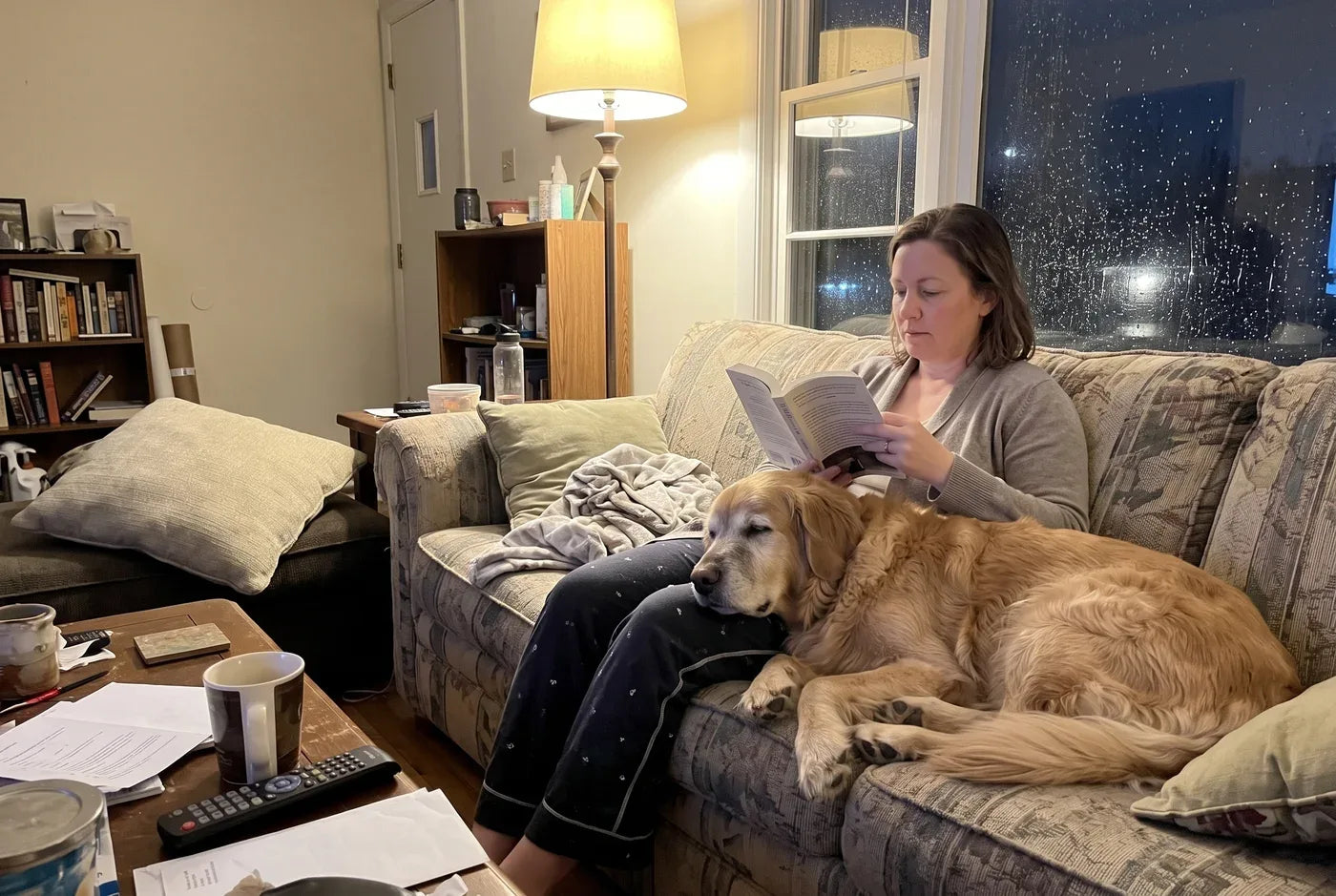 Photographic: a woman on a couch reading under a lamp during a rainstorm, rain streaking the window behind her, a medium