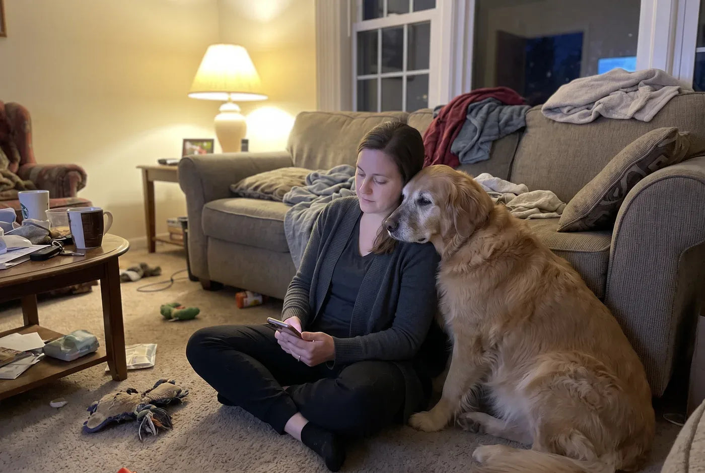 Photographic: a woman sitting on the floor with a phone, her medium-sized dog leaning against her shoulder, both relaxed